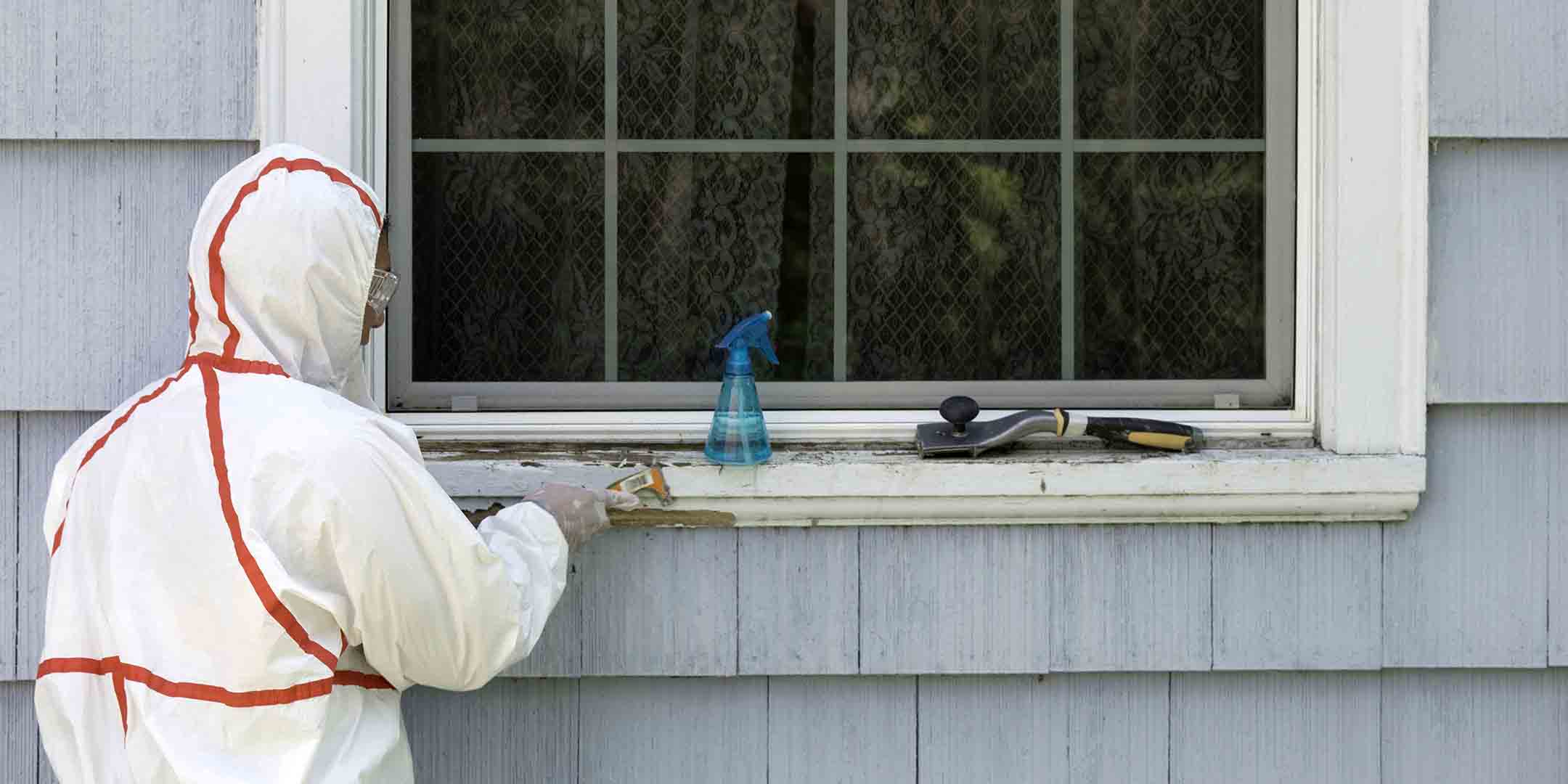 A house painter in a hazmat suit scrapes off dangerous lead paint from a window sill.